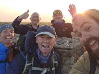 A group smile and wave at the camera at the final peak of the Welsh 3000s as the sun sets behind them